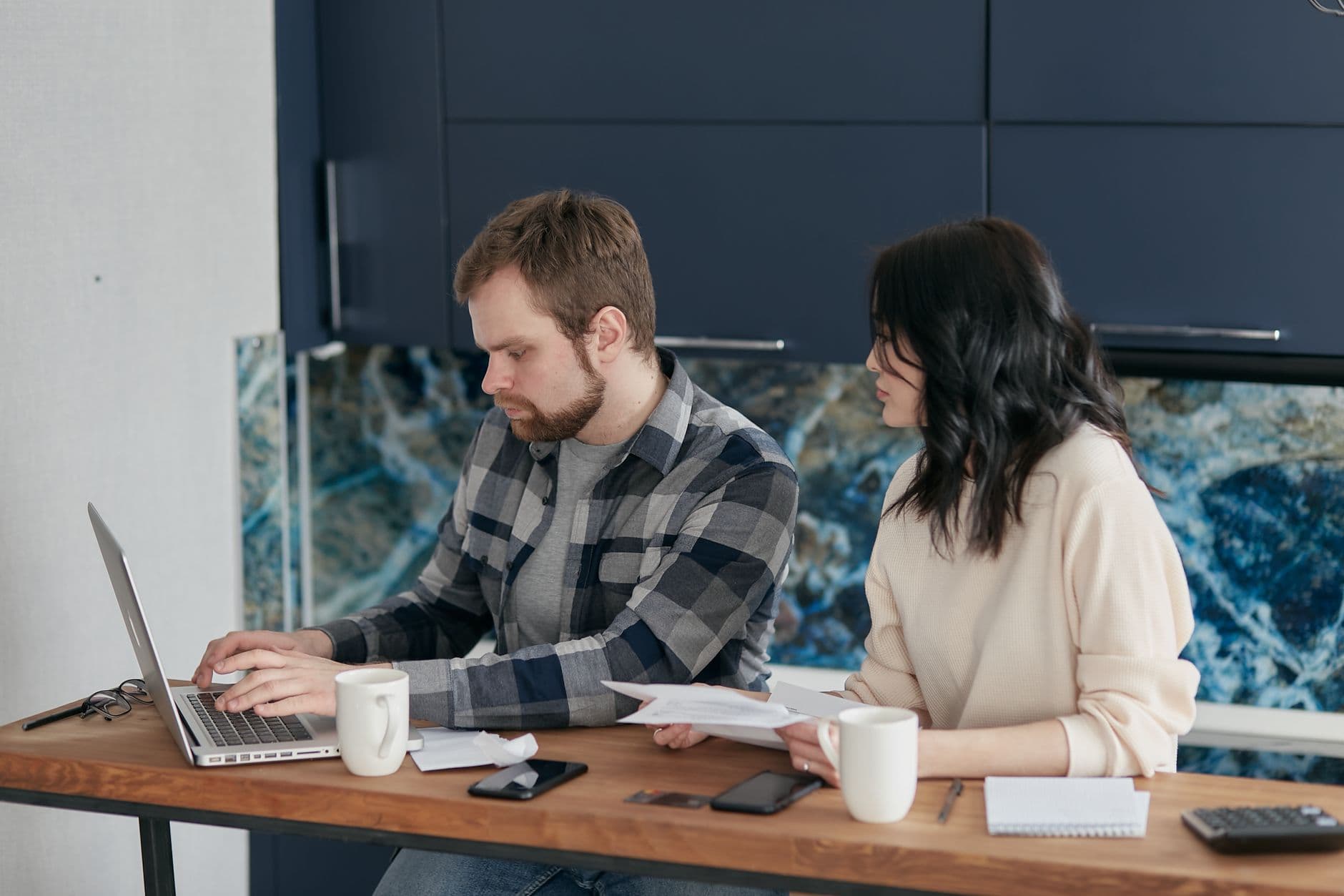 A couple sits at a kitchen table going through paperwork with a laptop.