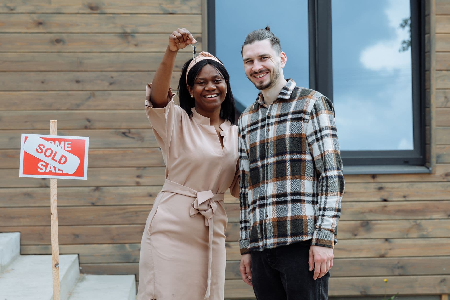 A couple stands in front of their new house next to a sold sign, holding up the key.
