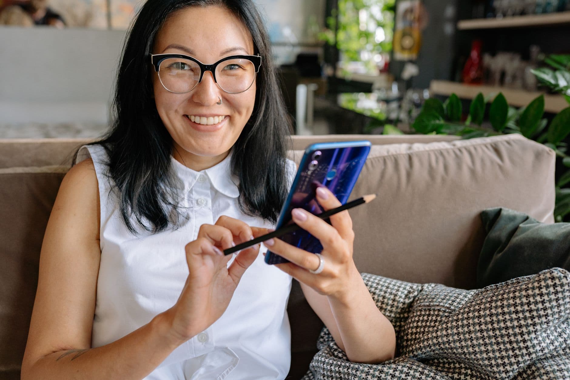 A smiling woman in glasses sits on a couch, looking up from her phone.