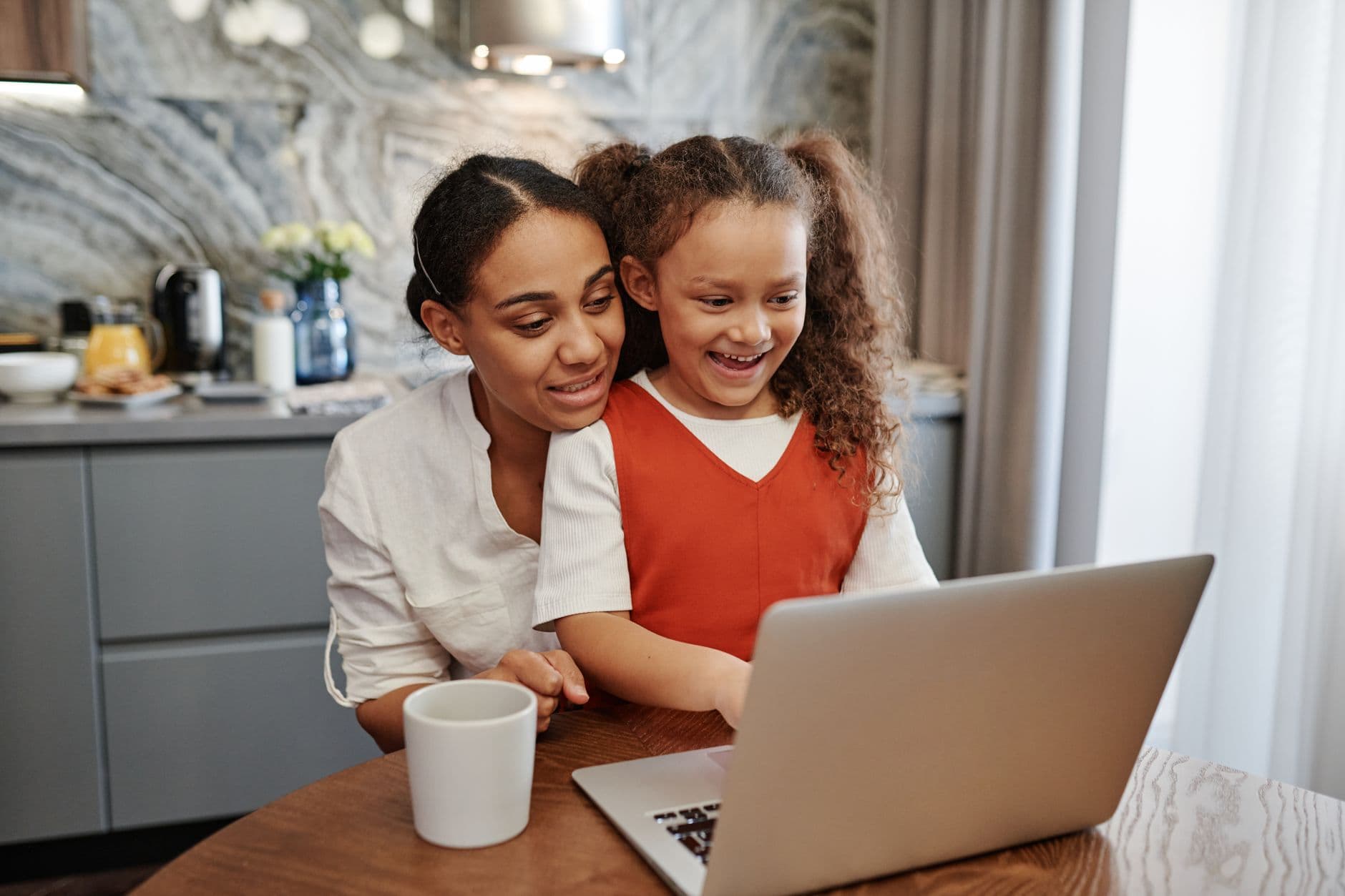 A mother and her young daughter smile at a laptop together in their kitchen.