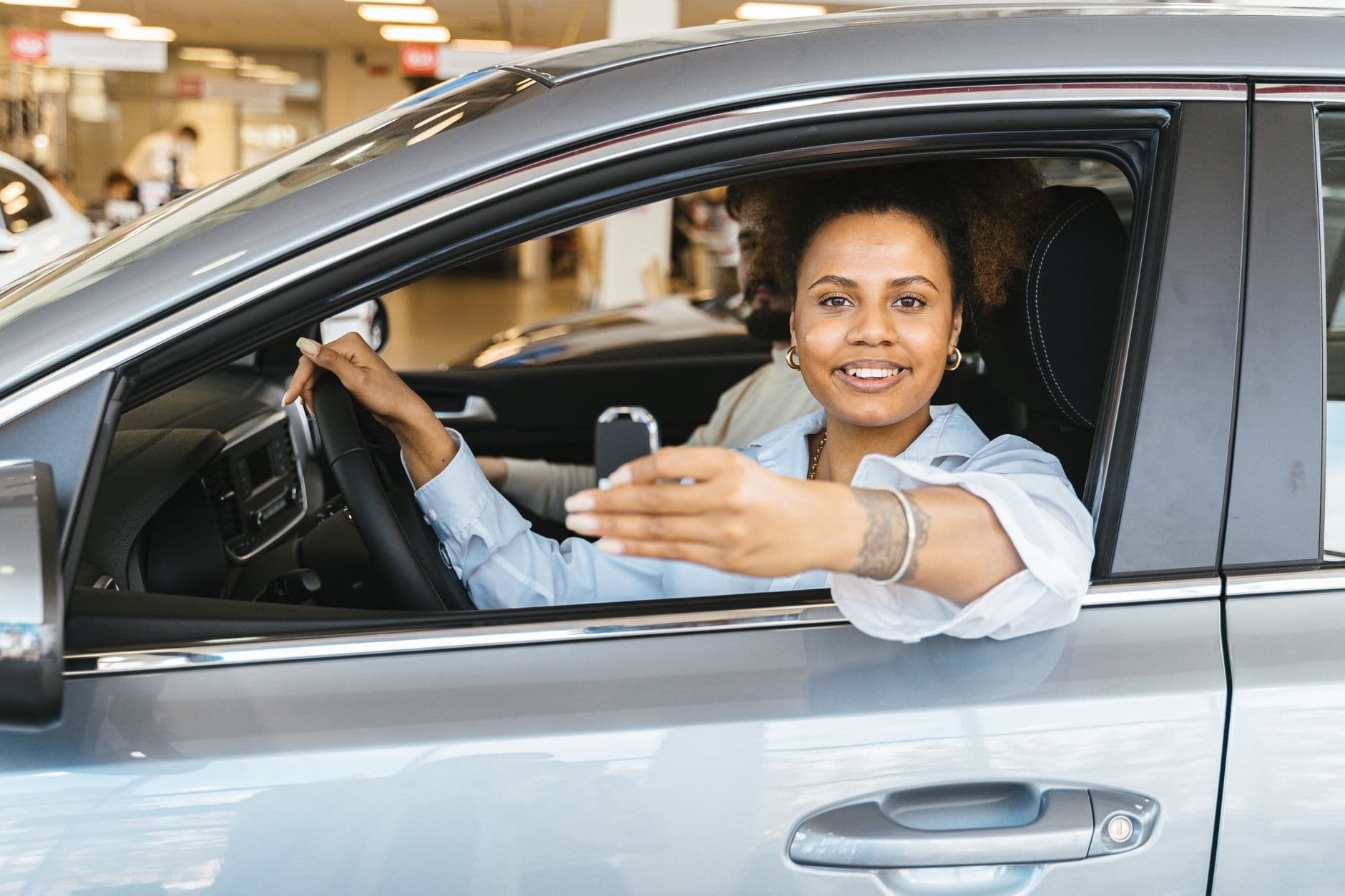 A woman sits in the driver's seat of a new car, smiling and holding up the key.